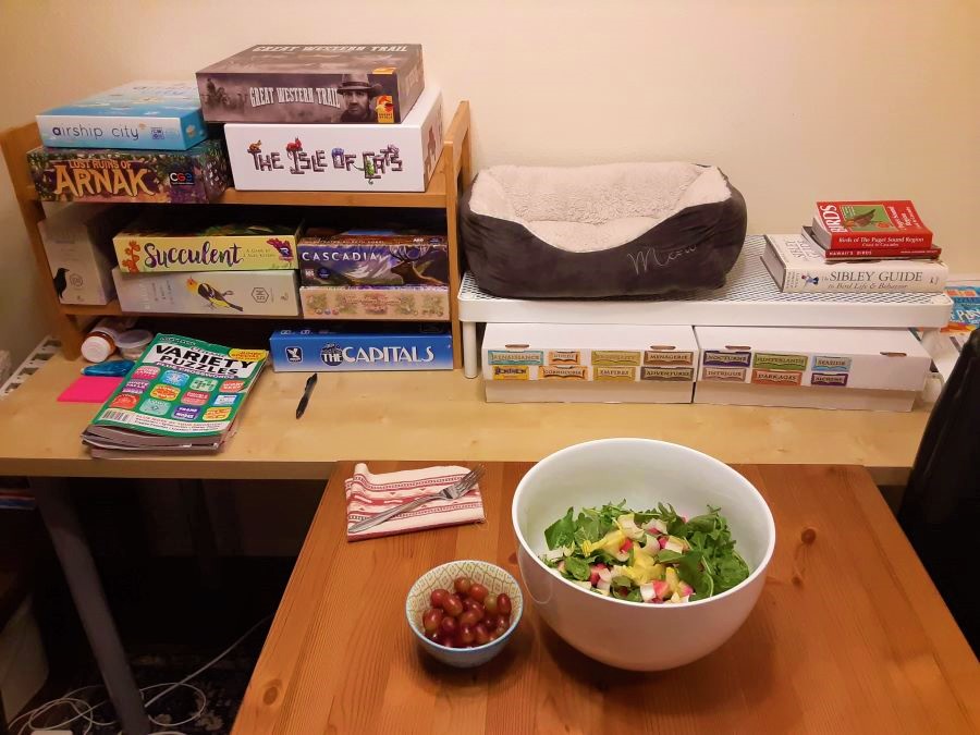 A dinner table with salad, fruit, puzzle magazines, books, and board games within arm's reach