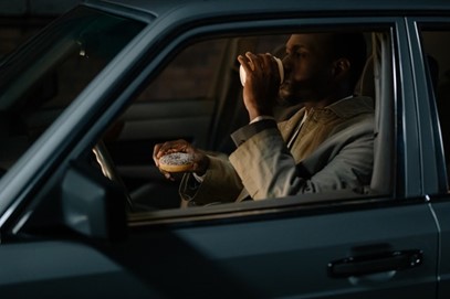 a man eating donut and drinking coffee in his car in the early morning