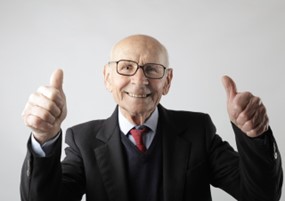 An elderly man in a suit smiling confidently with thumbs up