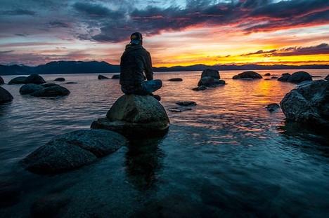 A man sitting on a rock on the shore, facing the sunset, with snowy mountains in the background.