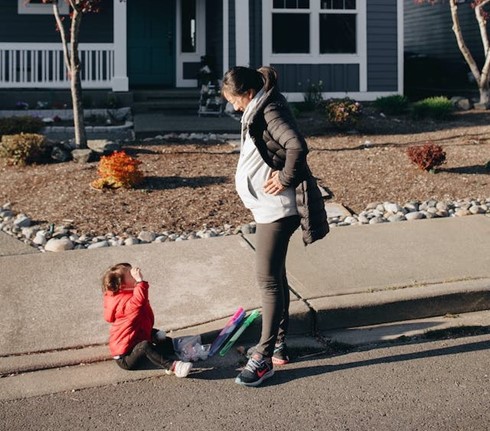 a parent looking down patiently at a child who has fallen on the sidewalk