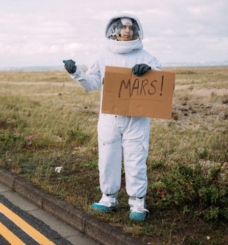 a man in an astronaut costume on the side of the road holding a cardboard sign that says "Mars!"