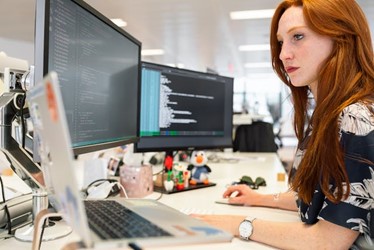 a busy woman working in an office with two monitors and a laptop in front of her