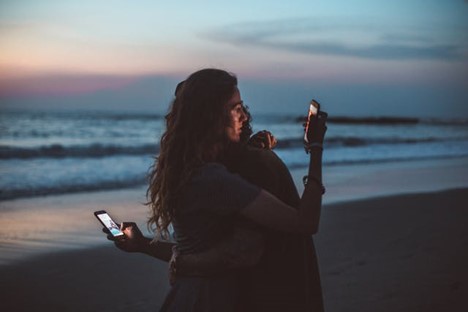 a couple on the beach at sunset, hugging, but looking at their phones